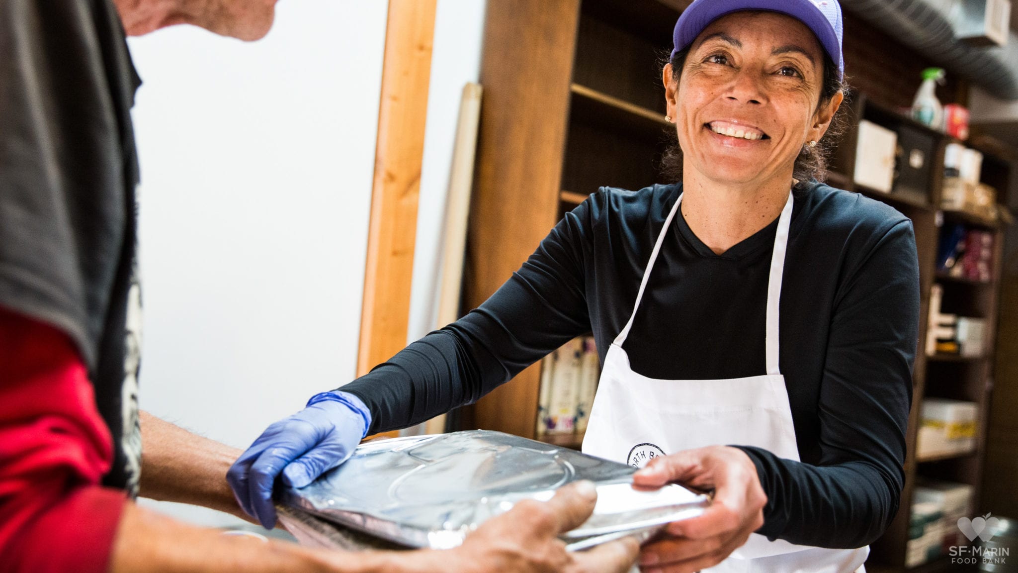 An SF Marin food bank volunteer in a white apron handing out a meal with a smile on her face